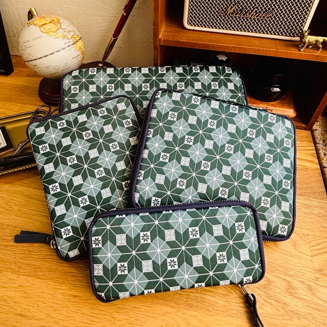 Three matching zippered pen cases with a green and white geometric star pattern and dark navy trim, arranged on a wooden tabletop with a globe, books, and a speaker in the background.