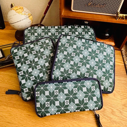 Three matching zippered pen cases with a green and white geometric star pattern and dark navy trim, arranged on a wooden tabletop with a globe, books, and a speaker in the background.