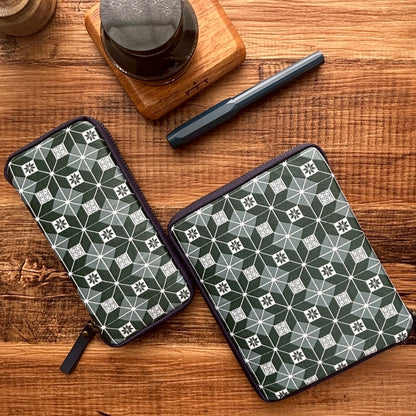 Two Kyoku Haku Restitch Emerald 10 Pen Cases from Piper Trading Co., featuring a green and white patterned design inspired by Siraya embroidery, rest on a wooden table alongside a black pen, an ink bottle, and a wooden coaster—ideal for elegantly storing your pen collection or using as a stylish fountain pen case.