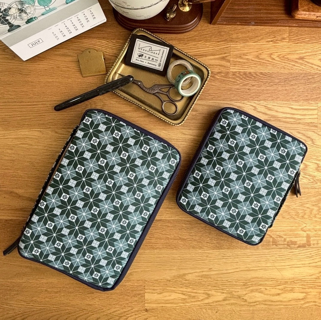The Kyoku Haku Restitch Emerald 10 Pen Case by Piper Trading Co., featuring green geometric patterns inspired by Siraya embroidery, sits on a wooden surface. Nearby are scissors, a gold tray with washi tape, a pen, and a calendar in the corner—making this stylish case perfect for your fountain pens or prized pen collection storage.