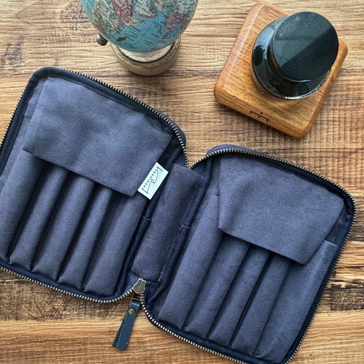 The Kyoku Haku Restitch Emerald 10 Pen Case by Piper Trading Co.—a navy blue zippered pen case with multiple compartments—is open on a wooden desk beside a small globe and a black ink bottle on a wooden base, making it an ideal choice for pen collection storage.