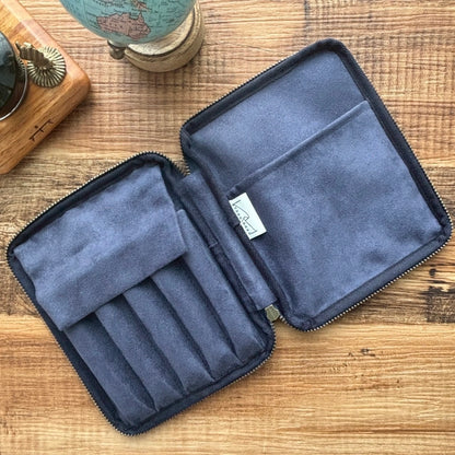 The Kyoku Haku Restitch Emerald 5 Pen Case by Piper Trading Co., featuring a soft fabric lining, secure zippered closure, and dedicated slots for pens, lies open on a wooden surface beside a small globe and a clock.