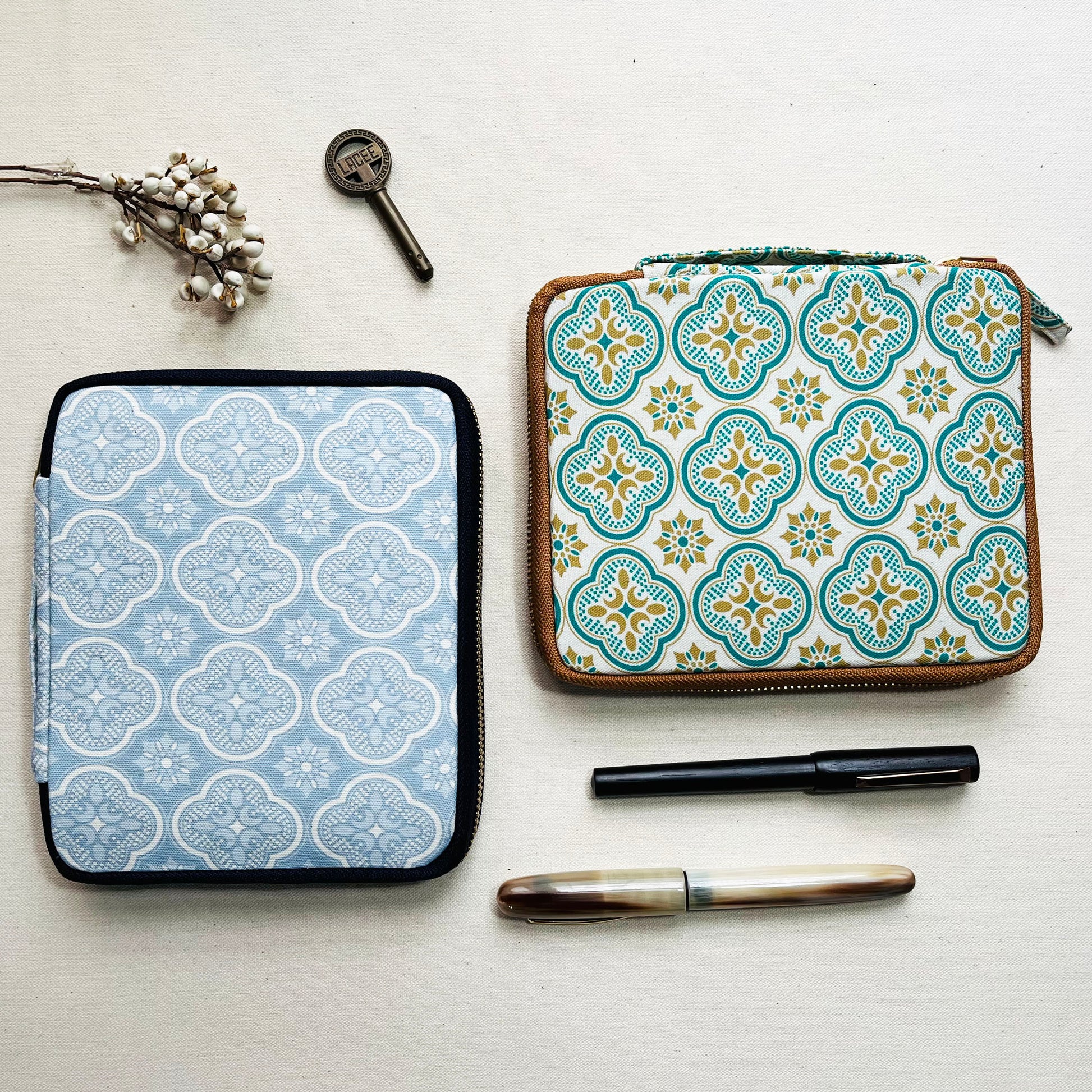 Two patterned Kyoku Haku Begonia Flowers 10 Pen Cases by Piper Trading Co.—one in blue and one in yellow-green—are placed beside a vintage key, dried flowers, and two pens on a neutral background. These handmade cases are perfect for stylishly storing your favorite writing instruments or fountain pens.