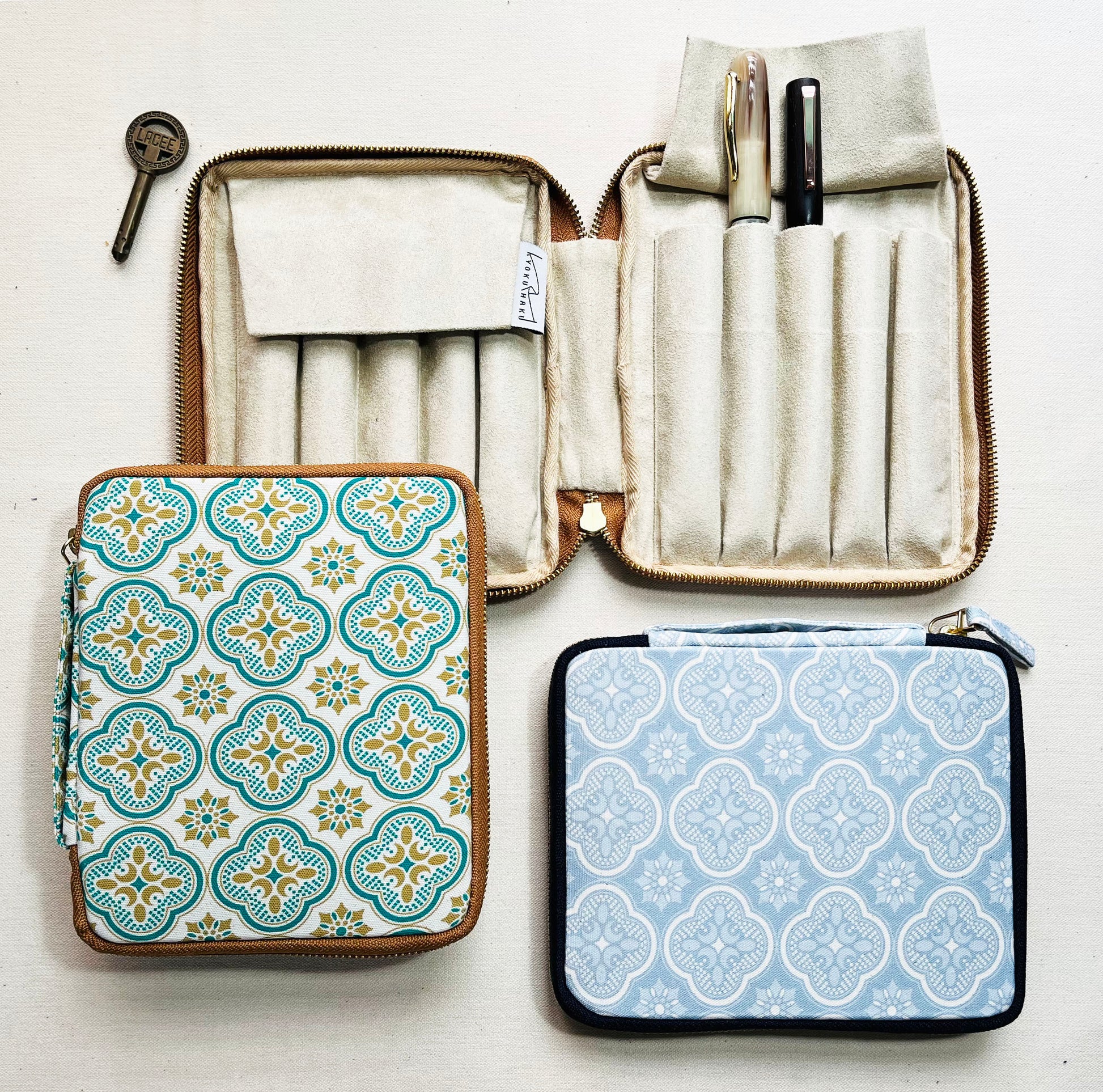 Three patterned zippered cases from Piper Trading Co. are displayed—a Kyoku Haku Begonia Flowers 10 Pen Case is open, showcasing fountain pens neatly arranged in holders, while two closed cases feature decorative tile-like designs. A small metal key rests beside the Kyoku Haku Begonia Flowers 10 Pen Case on a white surface.
