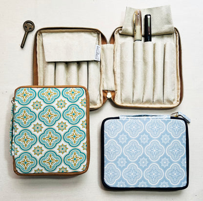 Three patterned zippered cases from Piper Trading Co. are displayed—a Kyoku Haku Begonia Flowers 10 Pen Case is open, showcasing fountain pens neatly arranged in holders, while two closed cases feature decorative tile-like designs. A small metal key rests beside the Kyoku Haku Begonia Flowers 10 Pen Case on a white surface.