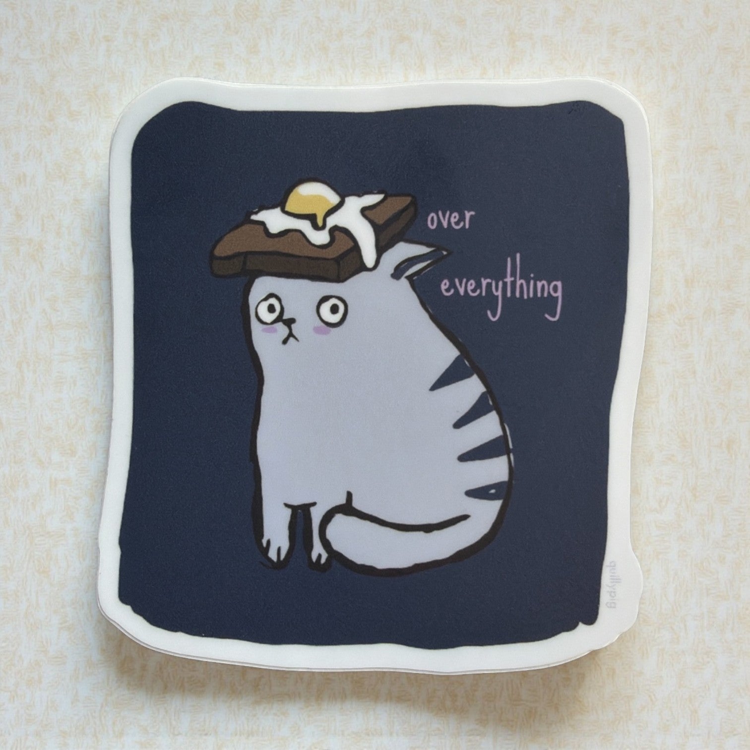 Piper Trading Co. sticker of a grumpy grey cat with a fried egg on toast on its head, with text 'over everything'.